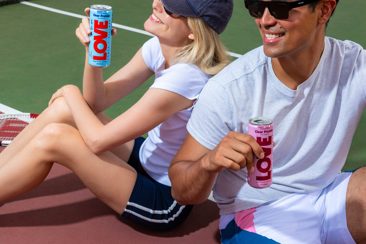 Two people relaxing on a tennis court with LOVE clear protein cans